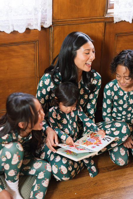 A woman and three children in Ivy City Co’s Baby So Soft Onesie in Very Merry Santa sit on a wooden floor by a window, smiling and reading a colorful picture book together.