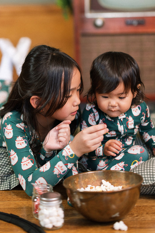 Two young children in matching Ivy City Co Baby So Soft Onesie in Very Merry Santa pajamas share popcorn from a large bowl, with a jar of marshmallows nearby amid festive decorations and a vintage TV.
