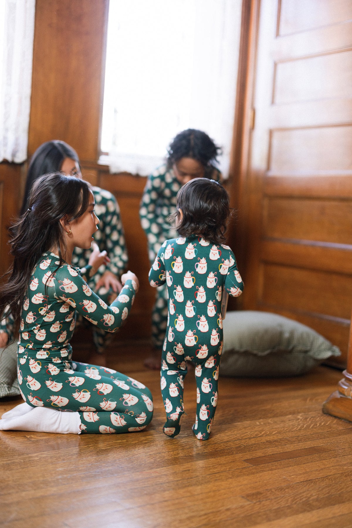 Children in Ivy City Co’s Baby So Soft Onesie in Very Merry Santa play on a hardwood floor near a pillow, while adults in cotton-modal knit outfits are blurred by the window and wooden wall.