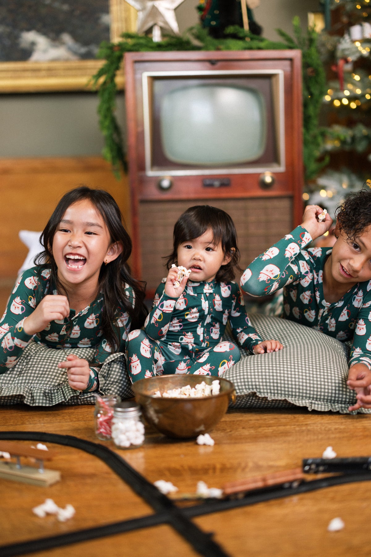 Three kids in Ivy City Co’s Baby So Soft Onesie in Very Merry Santa laugh by a vintage TV, eating popcorn. Pillows, a toy train track, and festive holiday décor complete the cozy holiday scene.