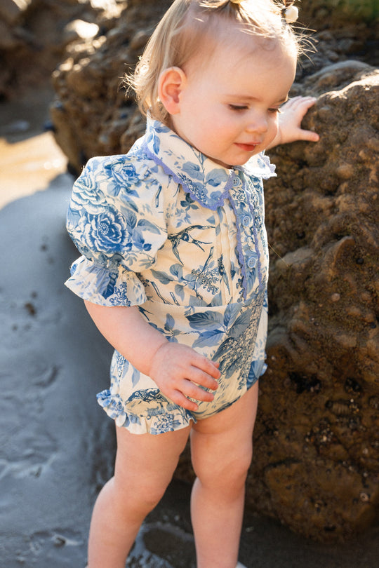 A toddler wearing the Ivy City Co Baby Sophia Romper in Blue Floral stands on the beach near a rock, sunlight highlighting their hair and outfit.