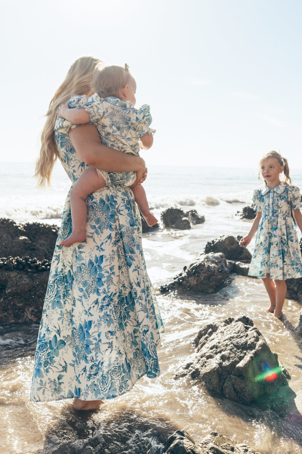 A woman holding a toddler stands barefoot on a rocky beach, both dressed in matching blue floral dresses. The toddler wears the Ivy City Co Baby Sophia Romper in Blue Floral, perfectly complementing her mother’s look. A young girl in a similar dress stands nearby in shallow water, sunlight shining brightly.