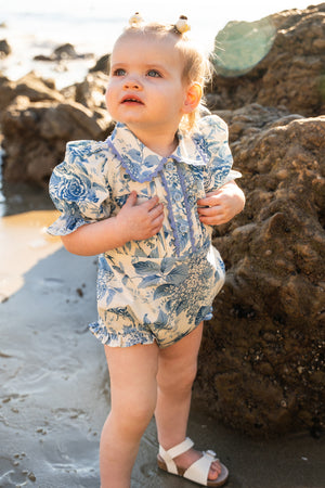 A toddler wearing the Ivy City Co Baby Sophia Romper in Blue Floral and white sandals stands on a sandy beach near rocks, looking up with sunlight shining on her face. Her hair is styled in two small buns.