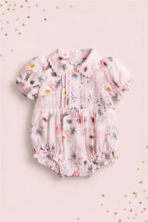 The Ivy City Co Baby Sophia Romper in Roselyn Pink features a floral pattern, lace chest accents, puffed short sleeves, and a rounded collar—perfect for mommy-and-me moments on a soft pink confetti-dotted background.