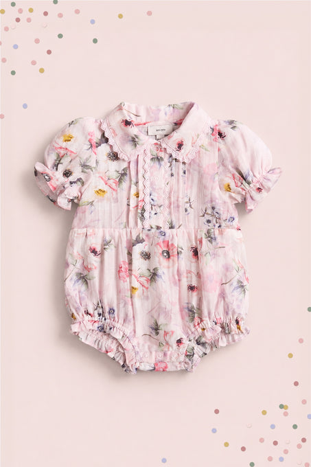 The Ivy City Co Baby Sophia Romper in Roselyn Pink features a floral pattern, lace chest accents, puffed short sleeves, and a rounded collar—perfect for mommy-and-me moments on a soft pink confetti-dotted background.