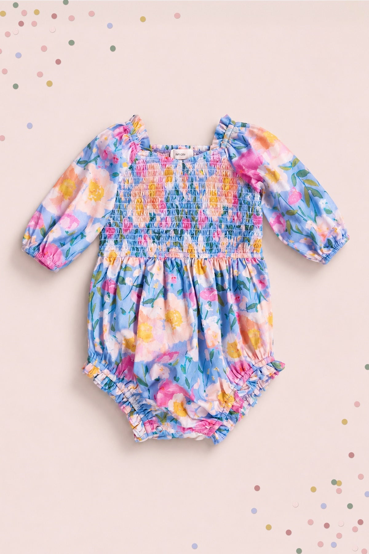 The Ivy City Co Baby Teagan Romper in Delia Blue features puffed sleeves, a smocked chest, and ruffled leg openings in a colorful floral print—ideal for a sweet mommy-and-me look.