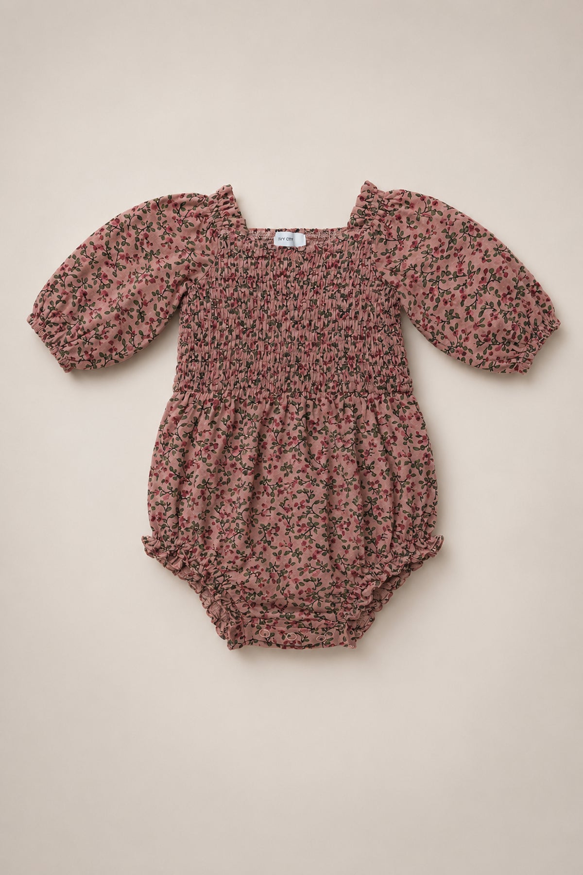The Ivy City Co Baby Teagan Romper in Mauve Woodland Floral is a charming cotton romper with puff sleeves and a smocked bodice, crafted from a pinkish-brown fabric adorned with a delicate small black floral print and shown laid flat on a light beige background.