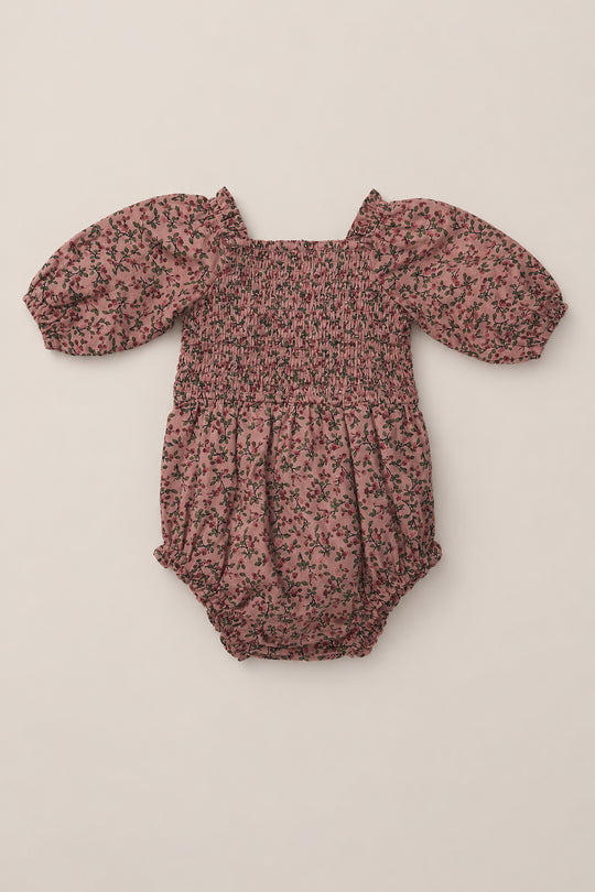 Introducing the Baby Teagan Romper in Mauve Woodland Floral from Ivy City Co—a soft cotton romper featuring a smocked bodice, puffed sleeves, and a charming black and green floral print on a mauve background. Designed with gathered elastic smocking at the chest and waist, this stylish piece lays beautifully flat for an adorable look.