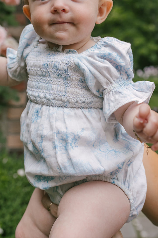 A baby, visible from the lower face down, is held outdoors against a blurred green background wearing the Ivy City Co Baby Valencia Romper—a white and light blue cotton piece with a smocked bodice.