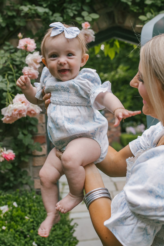 A smiling baby wearing the Ivy City Co Baby Valencia Romper with a smocked bodice and blue bow is held by a blonde woman; both match in light blue, posing together in a lush garden filled with pink roses.