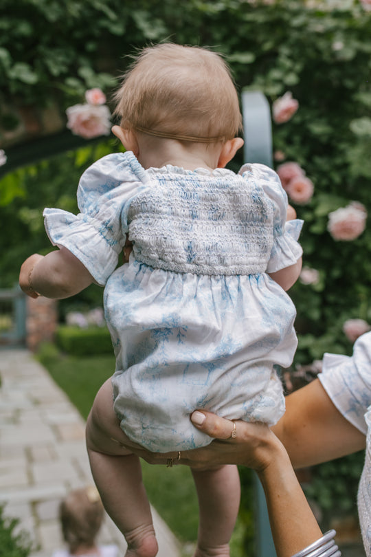 An adult holds up a baby wearing the Ivy City Co Baby Valencia Romper in light blue and white cotton. The baby’s back faces the camera, with pink roses and green foliage creating a lovely outdoor backdrop.