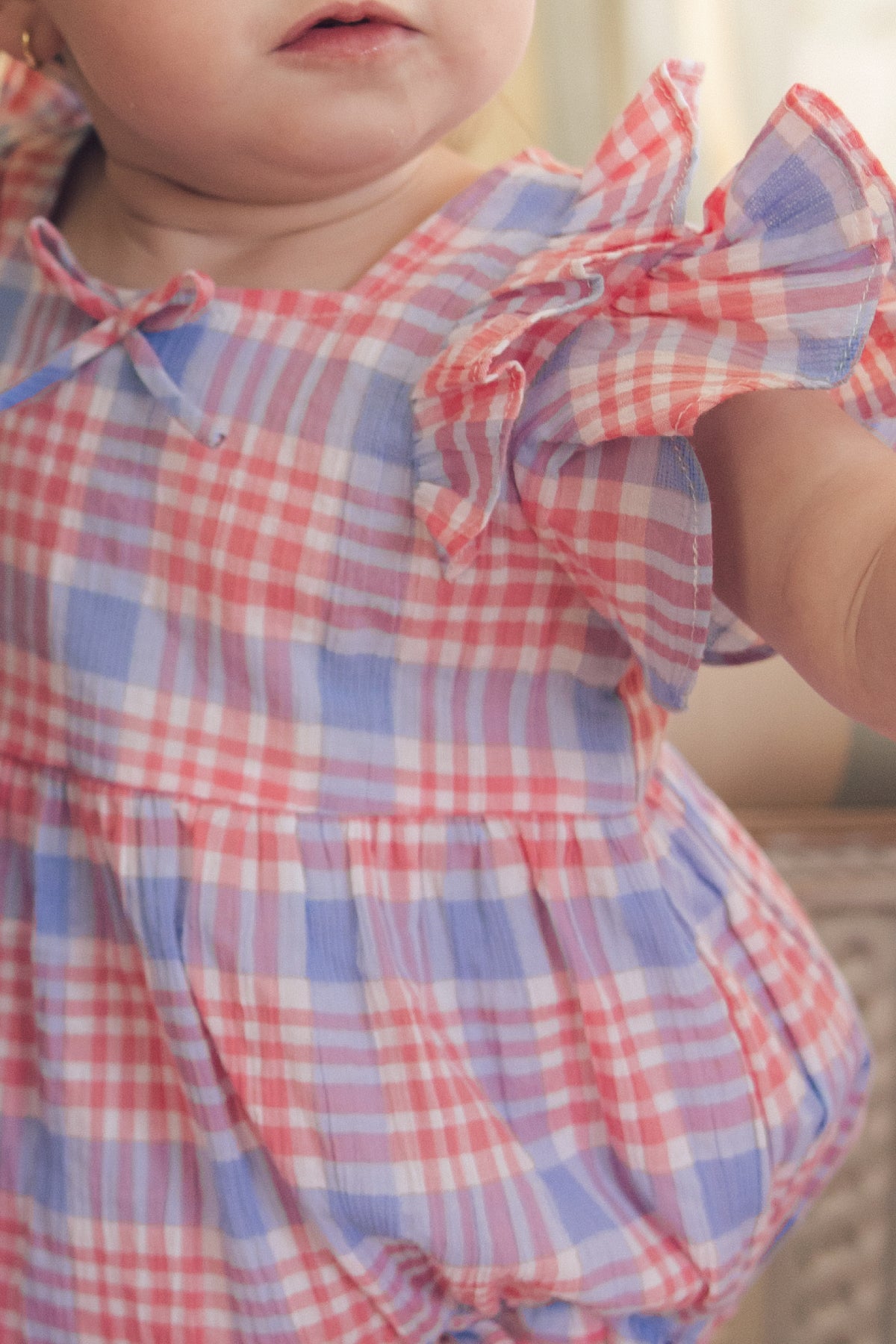 A close-up shows a baby wearing the Ivy City Co Baby Ana Romper in Blossom Plaid, featuring pastel pink, blue, and white with ruffled sleeves. The baby's face is partially visible against a softly blurred background.