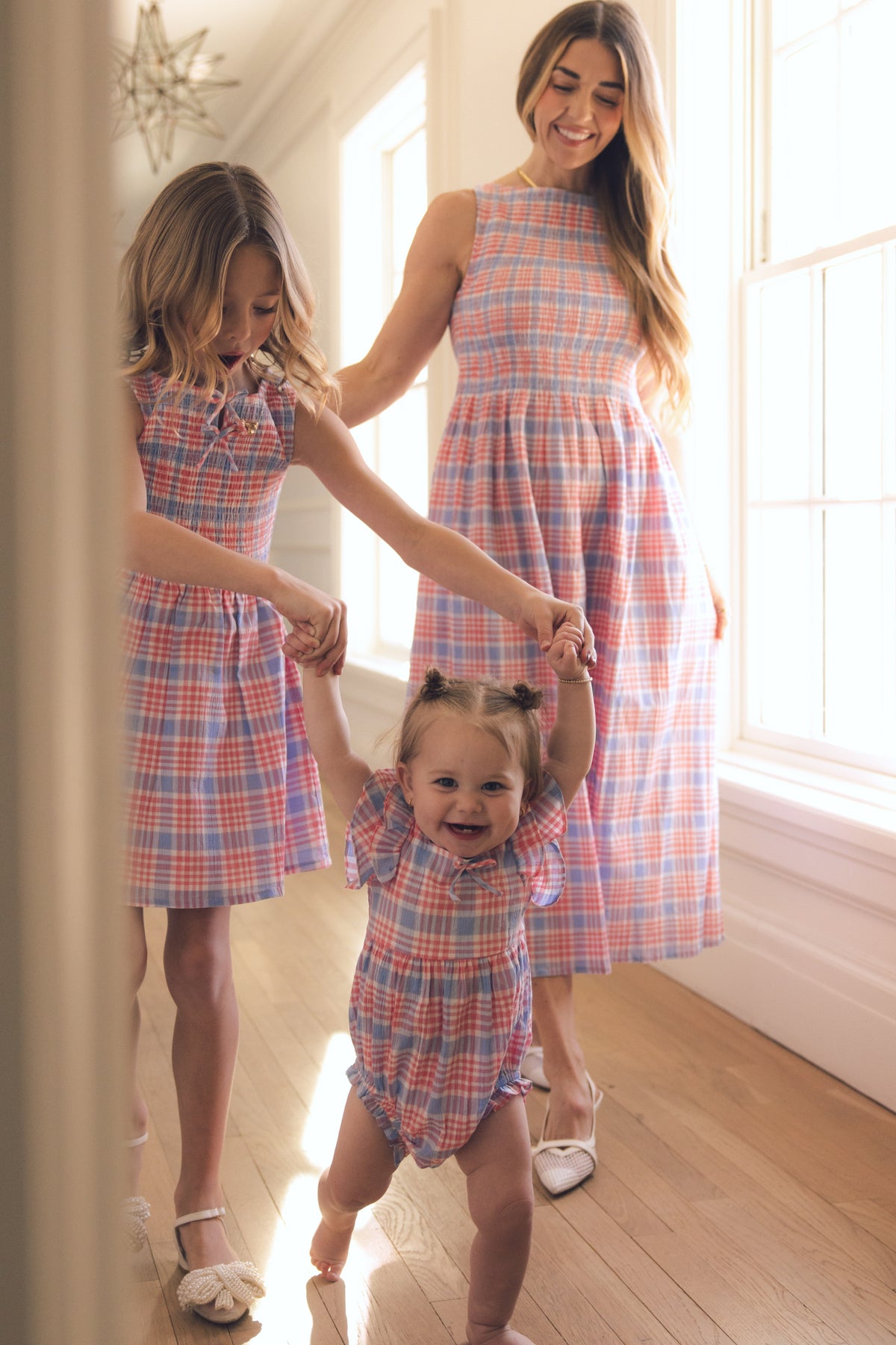 A woman, a young girl, and a toddler walk hand-in-hand in a sunlit hallway, all wearing Ivy City Co's blossom plaid outfits. The toddler beams with joy in the Baby Ana Romper in Blossom Plaid, supported by the others.
