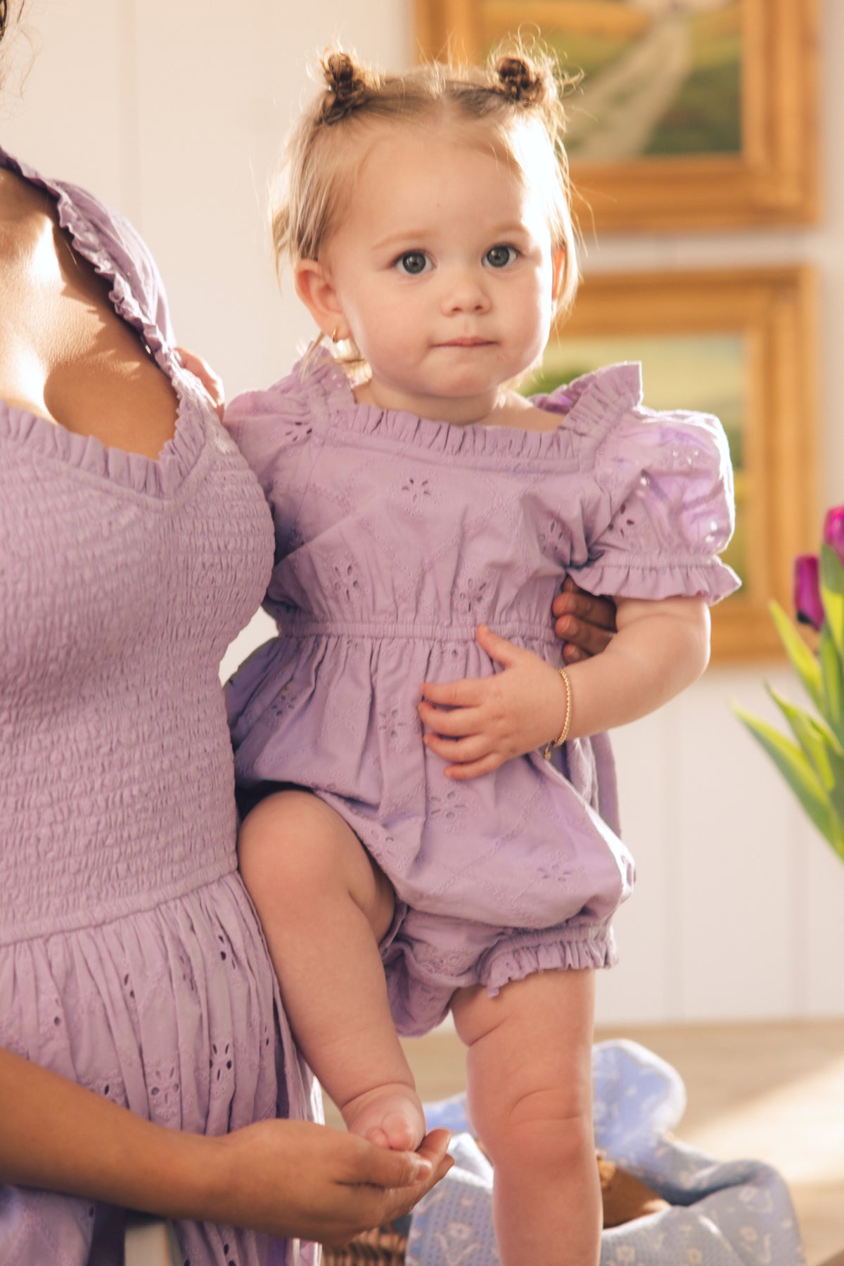 A baby with light hair in two buns sits on a woman’s hip. Both wear Ivy City Co’s Baby Emmi Romper in Lilac—an ideal spring outfit. The softly lit scene features blurred paintings and flowers in the background.