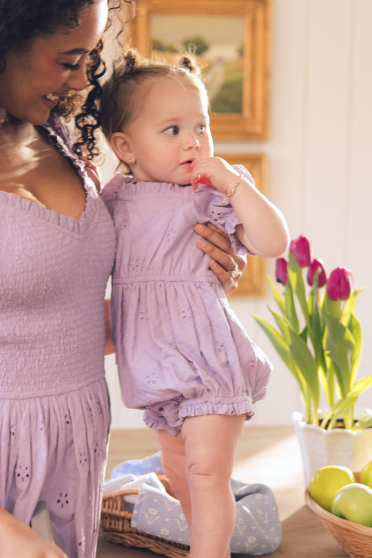 A woman in a purple outfit holds a baby dressed in the adorable Baby Emmi Romper in Lilac by Ivy City Co, standing beside a table adorned with a vase of purple tulips and a bowl of green apples. The baby looks to the side, touching their mouth—perfectly styled for spring in this charming Ivy City Co piece.