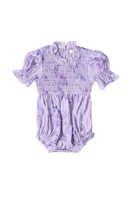 The Baby Flynn Romper in Lavender Aquarelle Fleur by Ivy City Co is a lavender floral romper with short puff sleeves, ruffled edges, smocked chest, elastic leg openings, and a subtle watercolor print, shown laid flat on a white background.