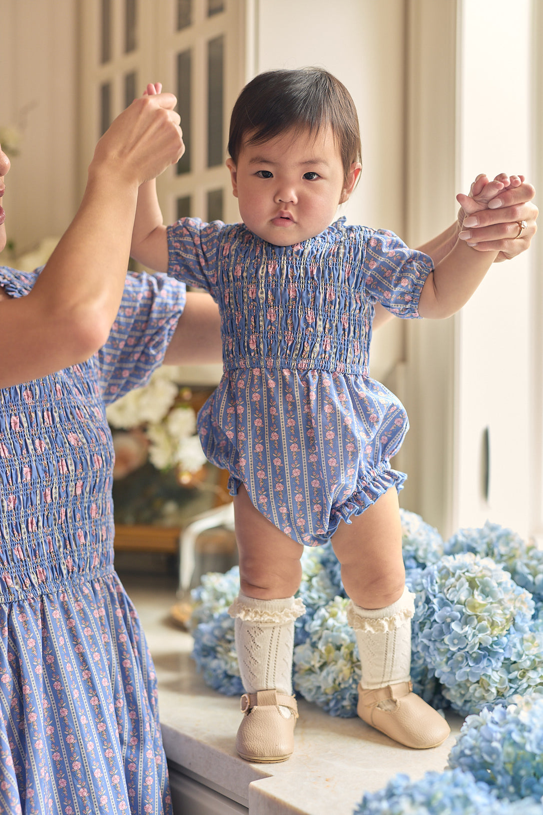 A baby wearing the Ivy City Co Baby Mariah Romper in Linear Print with white boots stands on a counter, holding an adult’s hands for support. The adult wears a matching blue floral outfit, with blue hydrangeas blooming in the background.