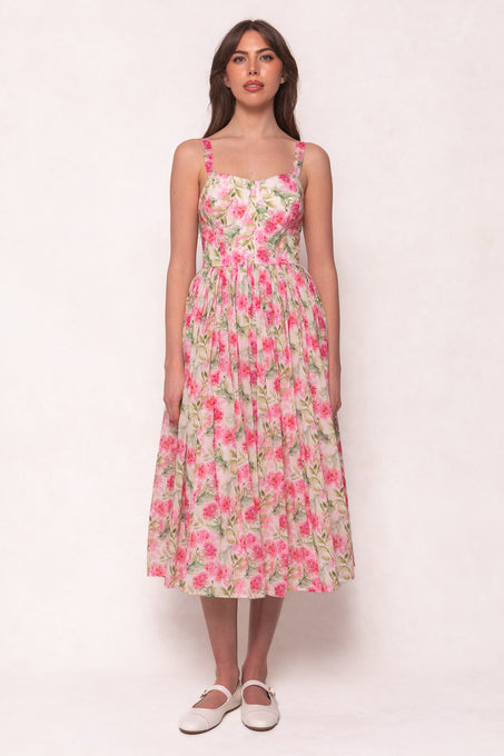 A woman with long brown hair and neutral makeup wears the Ivy City Co Ballerina Tank Dress in Pink Floral, featuring a pleated, midi-length skirt, against a light background. She completes the look with white flat shoes.