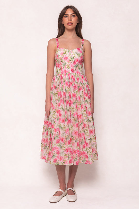 A woman with long brown hair and neutral makeup wears the Ivy City Co Ballerina Tank Dress in Pink Floral, featuring a pleated, midi-length skirt, against a light background. She completes the look with white flat shoes.