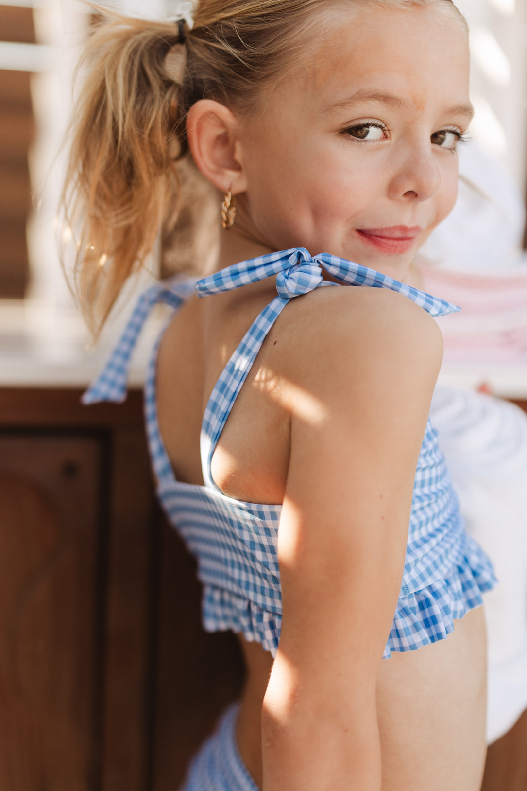 A young girl with blonde hair in a ponytail smiles over her shoulder, wearing the Ivy City Co Mini Brandi Bandeau Top in Blakely Gingham, standing in sunlight with a wooden background behind her.