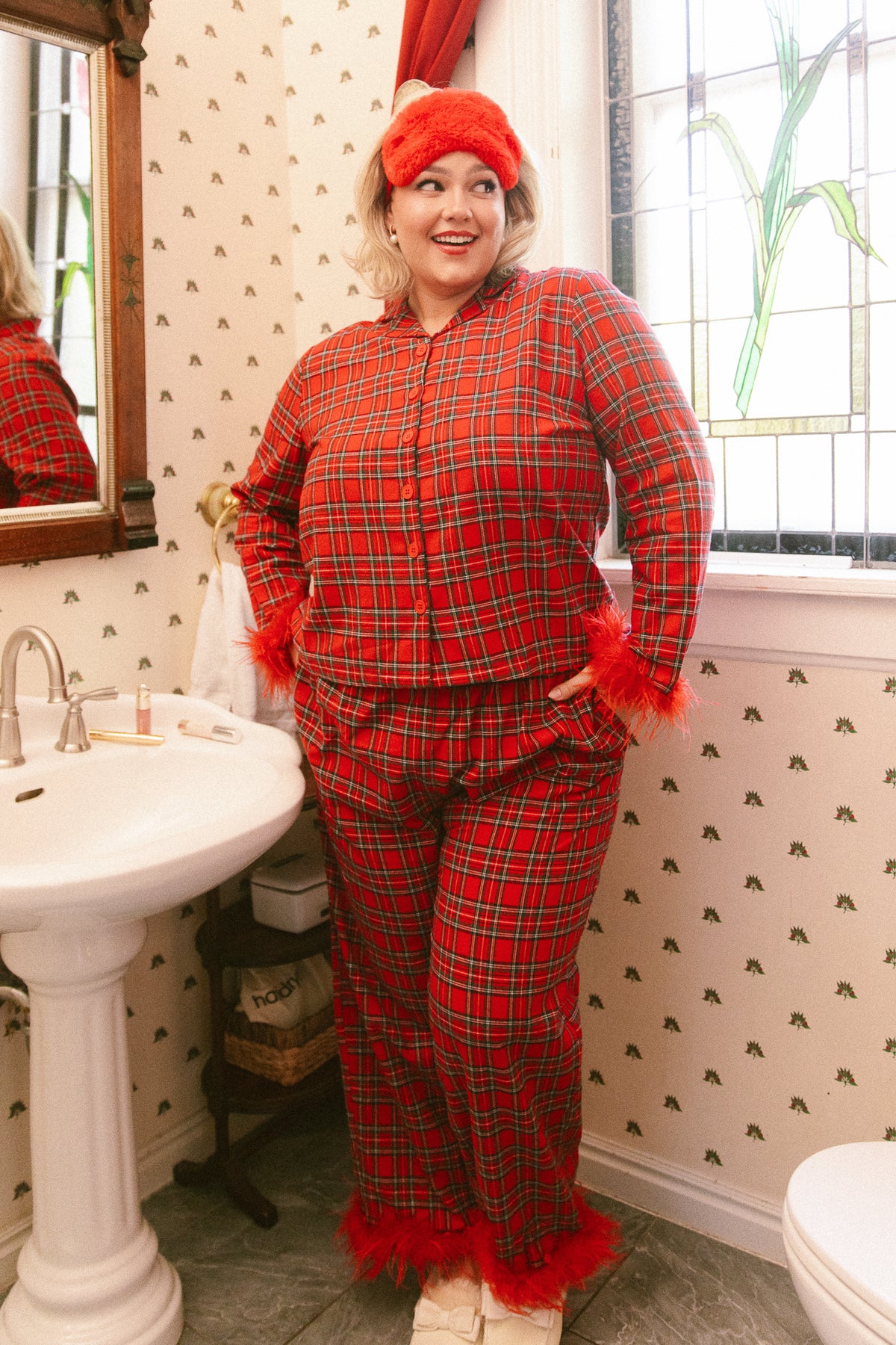 A woman standing in a bathroom wearing a Benjamin Pajama Set in Holiday Plaid, featuring red feather trim on the sleeves and pant cuffs, with a matching red eye mask on her head.