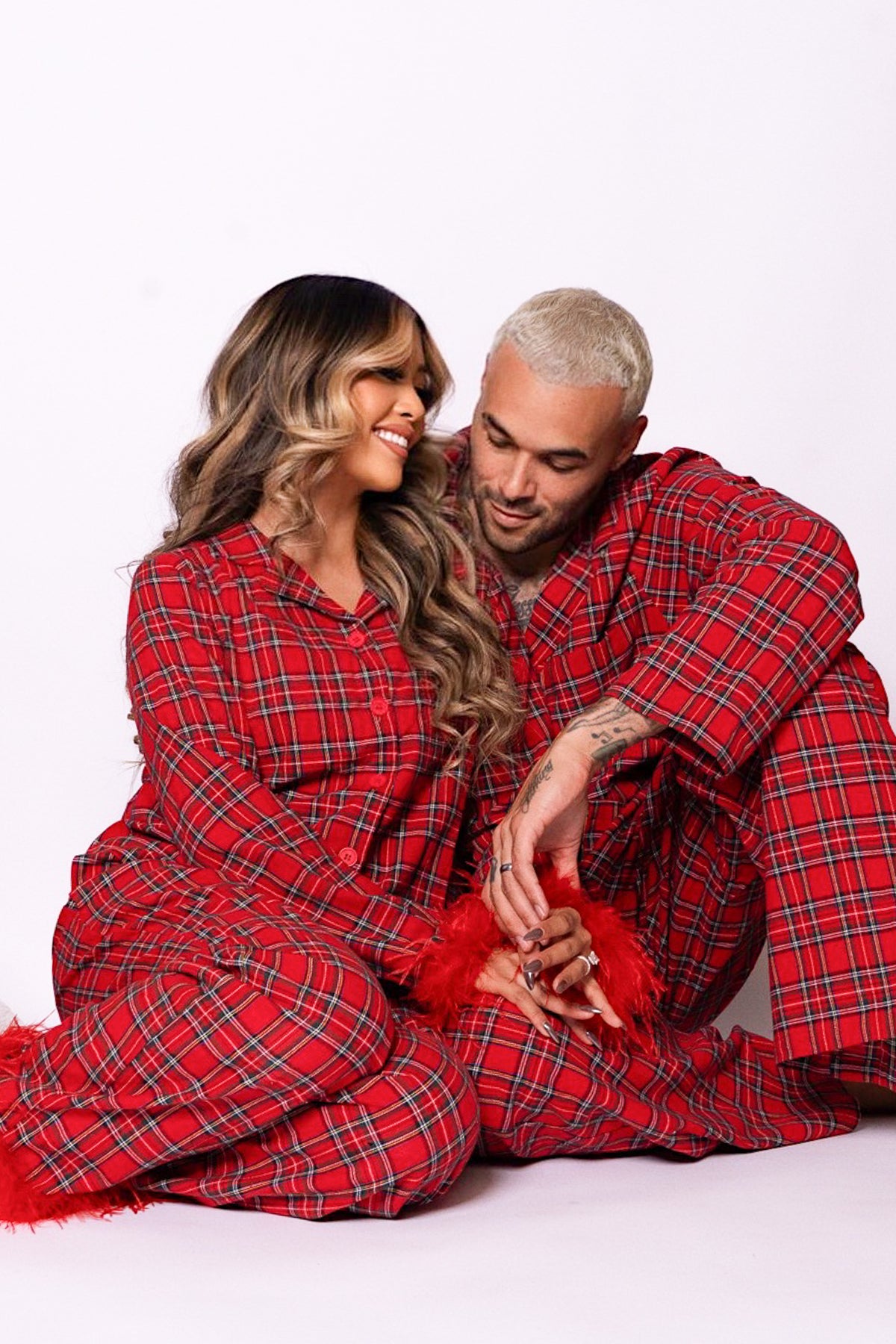 A man and woman sitting together wearing matching Benjamin Pajama Set in Holiday Plaid button-downs with feather trim cuffs.