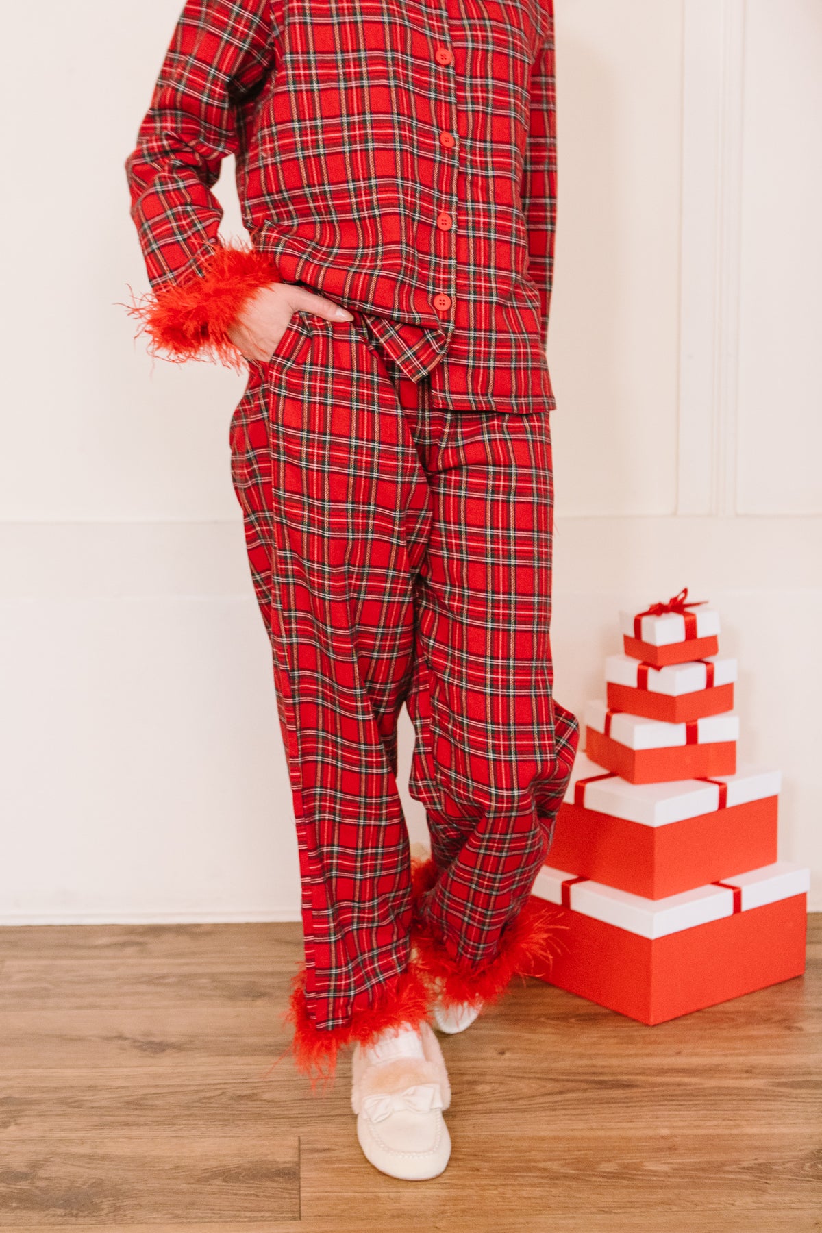 A person wearing a red Benjamin Pajama Set in Holiday Plaid with feather trim at the cuffs and ankles, standing next to a stack of red and white gift boxes.