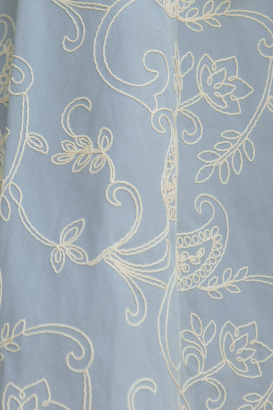 The Bennett Dress in Cream Embroidery by Ivy City Co features light blue fabric adorned with intricate cream floral and vine embroidery, adding elegance and a graceful swirl of flowers and leaves to its textured midi silhouette.