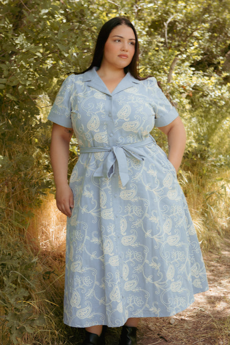 A woman wearing the Ivy City Co Bennett Dress in Cream Embroidery—a nursing-friendly, chambray denim midi dress with a white paisley pattern—stands outdoors on a sunlit dirt path amid green foliage.