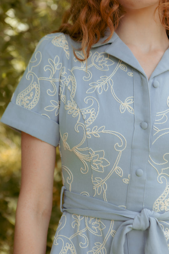A close-up of a person wearing the Ivy City Co Bennett Dress in Cream Embroidery, featuring short sleeves, white floral embroidery, a collared neckline, nursing-friendly front buttons, and a tied waist belt. Blurred greenery background.