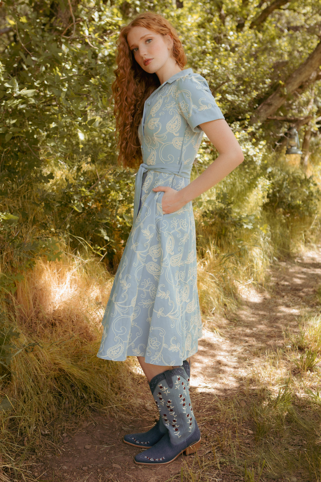 A young woman with long red curly hair stands outdoors on a sunlit path, wearing the Ivy City Co Bennett Dress in Cream Embroidery, paired with blue cowboy boots decorated with silver studs.