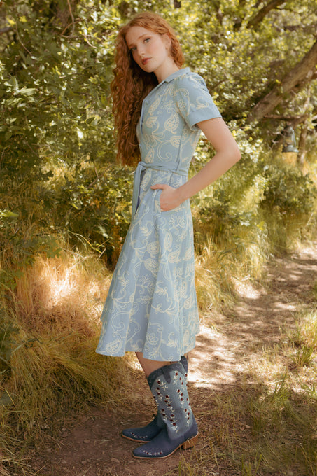 A young woman with long red curly hair stands outdoors on a sunlit path, wearing the Ivy City Co Bennett Dress in Cream Embroidery, paired with blue cowboy boots decorated with silver studs.