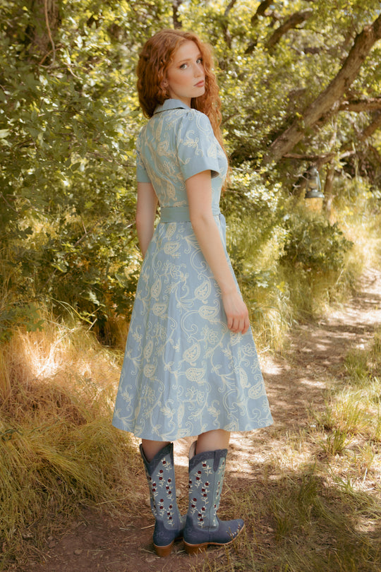A woman with long red hair stands on a sunlit forest path, wearing the Bennett Dress in Cream Embroidery by Ivy City Co. She looks over her shoulder toward the camera.
