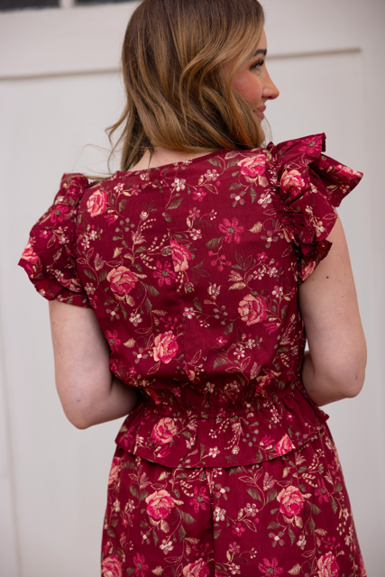 A woman with wavy light brown hair stands sideways, showcasing the back of the Berlin Dress in Red by Ivy City Co—a feminine maroon floral dress with ruffled sleeves and cinched waist—against a plain, light-colored wall.