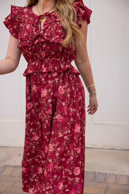A woman stands on a brick sidewalk in the Ivy City Co Berlin Dress in Red, a feminine burgundy floral midi dress with ruffles. Her long, loose waves and jewelry complete the look. Her face is not visible.