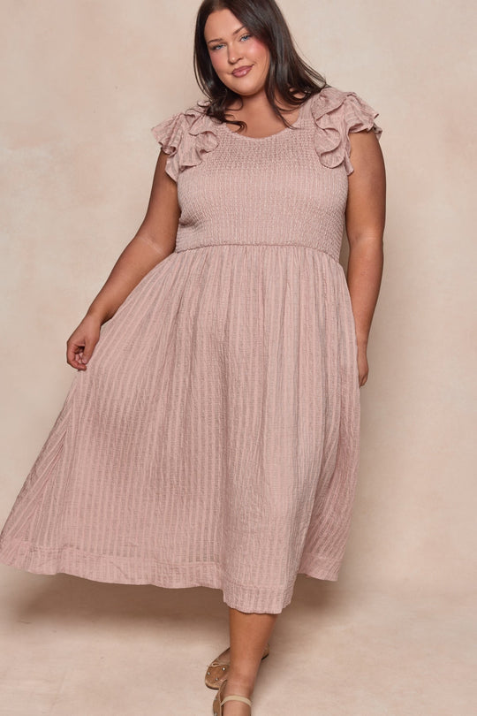 A woman wearing the Ivy City Co Hattie Dress in Pink Stripes—a light pink, short-sleeved, ruffled midi dress with a smocked bodice—stands against a neutral background, smiling and holding the skirt to the side. This charming maternity-friendly style offers both comfort and elegance.