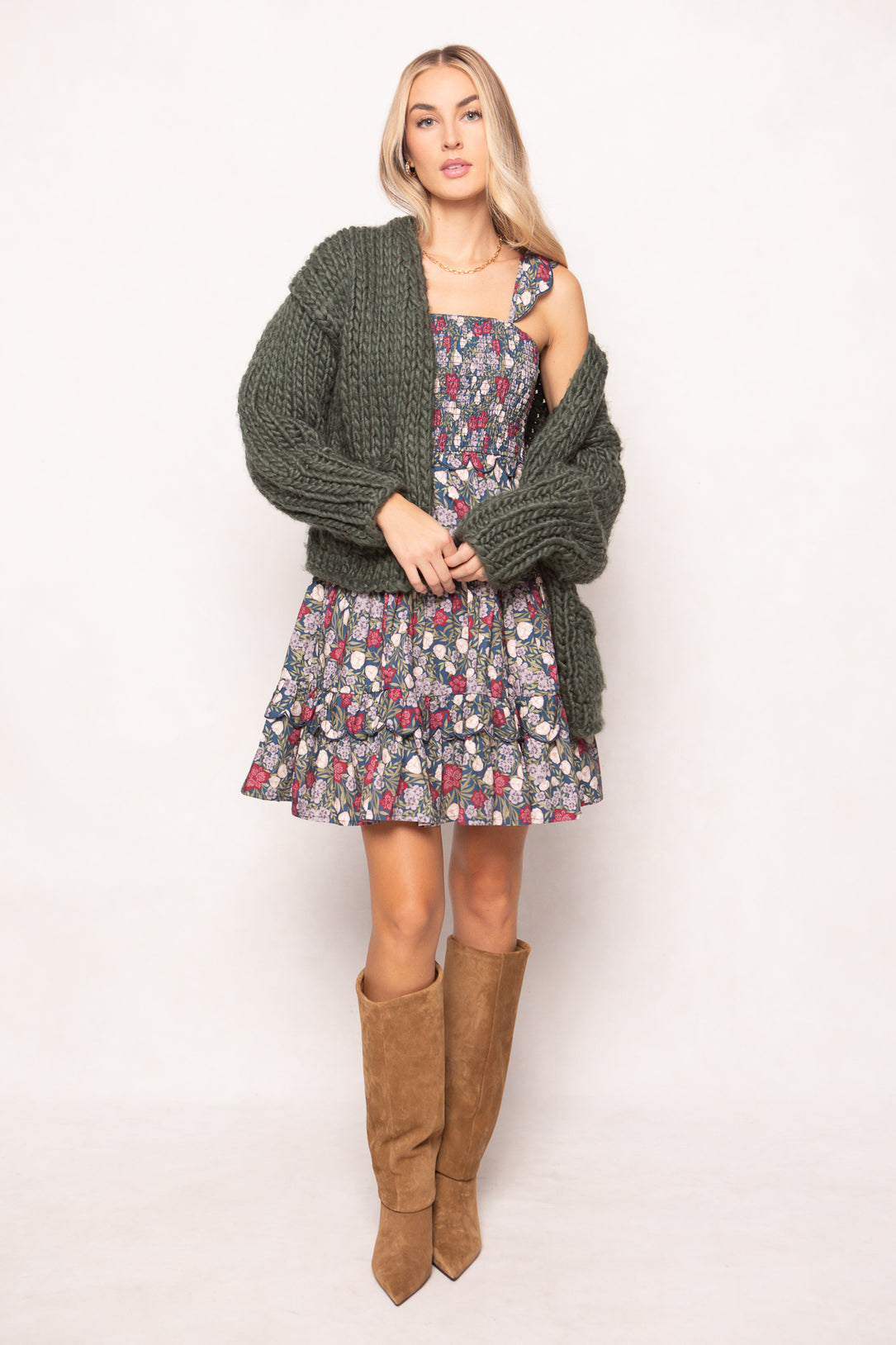 A woman stands against a white background wearing Ivy City Co's Blossom Dress in Navy Meadow, paired with an oversized green knit cardigan and tan suede boots—a stylish, nursing-friendly look perfect for moms on the go.