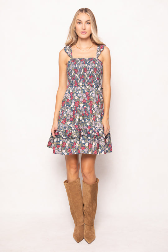 A woman stands against a white background, wearing the Ivy City Co Blossom Dress in Navy Meadow, a sleeveless floral design with tied shoulder straps and brown knee-high boots. This maternity friendly style suits her long blond hair and neutral expression.