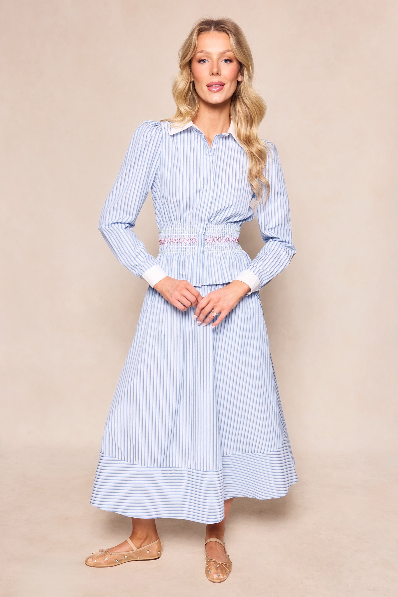 A woman with long blonde hair stands against a beige background, wearing the Ivy City Co Bluebell Top in blue and white stripes styled as a dress, featuring a smocked waistband and flared mid-calf skirt. She finishes the look with tan flat shoes.