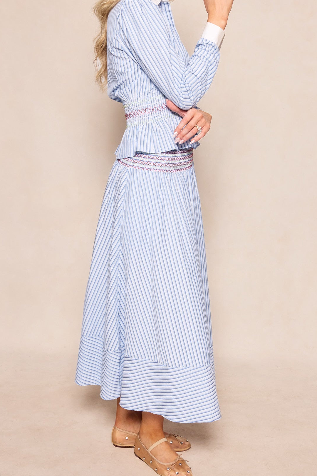 A woman stands sideways wearing Ivy City Co’s Bluebell Skirt in Blue & White Stripes, styled with a maternity-friendly, long-sleeved dress, smocked waistband, and clear studded sandals. Her light hair is loose, hands gently clasped.