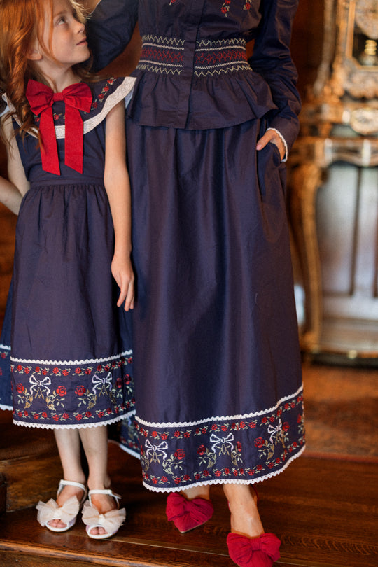 Both the child and adult wear matching navy blue outfits with red and white embroidery from Ivy City Co. The child, dressed in the Bluebell Skirt in Sapphire Rosette Embroidery, looks up at the adult as they stand together on a wooden floor. Only the lower halves of their faces and bodies are visible.