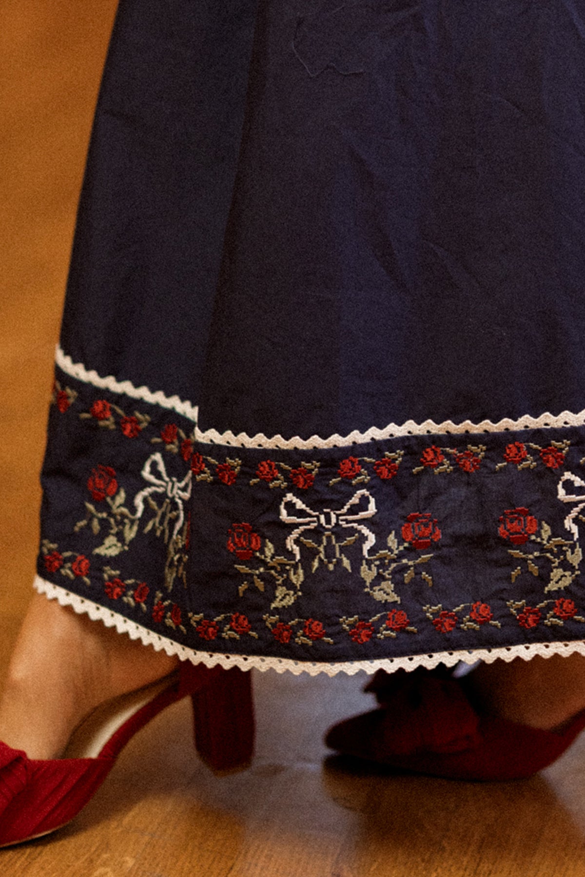 A close-up of the Ivy City Co Bluebell Skirt in Sapphire Rosette Embroidery, featuring intricate floral lace detailing and trim in a classic A-line cotton silhouette, paired with red heeled shoes on a wooden floor—perfect for a festive girls’ holiday ensemble.