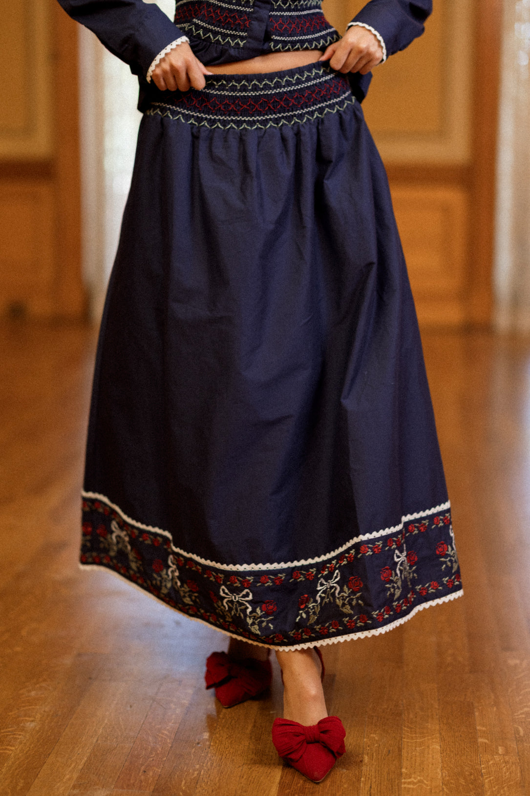 A person wearing the Ivy City Co Bluebell Skirt in Sapphire Rosette Embroidery, paired with red shoes with bows, standing on a wooden floor.