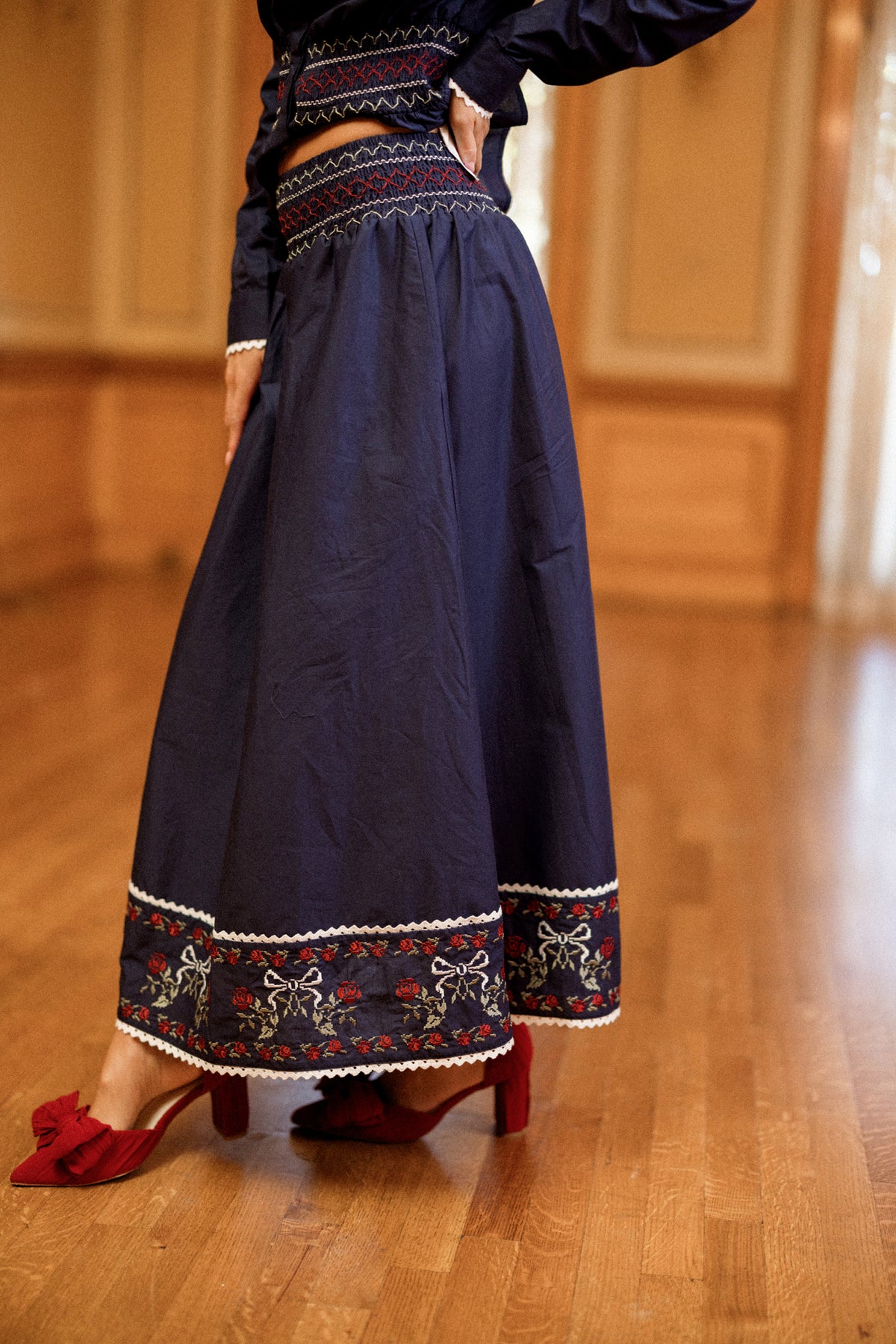 A woman wearing the Ivy City Co Bluebell Skirt in Sapphire Rosette Embroidery, an A-line cotton skirt in navy with embroidered patterns near the hem, paired with red heeled shoes. The setting is an indoor wooden floor with tan walls.