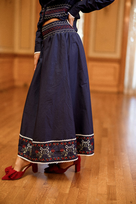 A woman wearing the Ivy City Co Bluebell Skirt in Sapphire Rosette Embroidery, an A-line cotton skirt in navy with embroidered patterns near the hem, paired with red heeled shoes. The setting is an indoor wooden floor with tan walls.