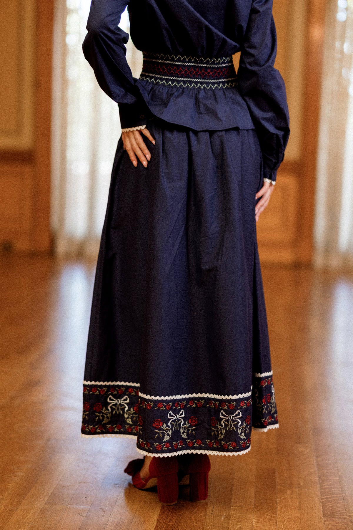 A person stands on a wooden floor with hands on hips, partially turned away from the camera and wearing Ivy City Co’s Bluebell Skirt in Sapphire Rosette Embroidery—a navy blue A-line cotton design featuring red and white floral embroidery along the hem and waist.