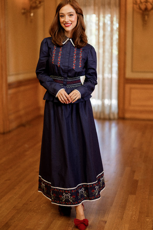 A woman stands indoors wearing the Ivy City Co Bluebell Skirt in Sapphire Rosette Embroidery, a long navy skirt adorned with red and white embroidered details reminiscent of lace. She pairs it with a white collared top and long sleeves, has brown hair, is smiling, and completes her outfit with red shoes. The background features wood paneling and sheer curtains.