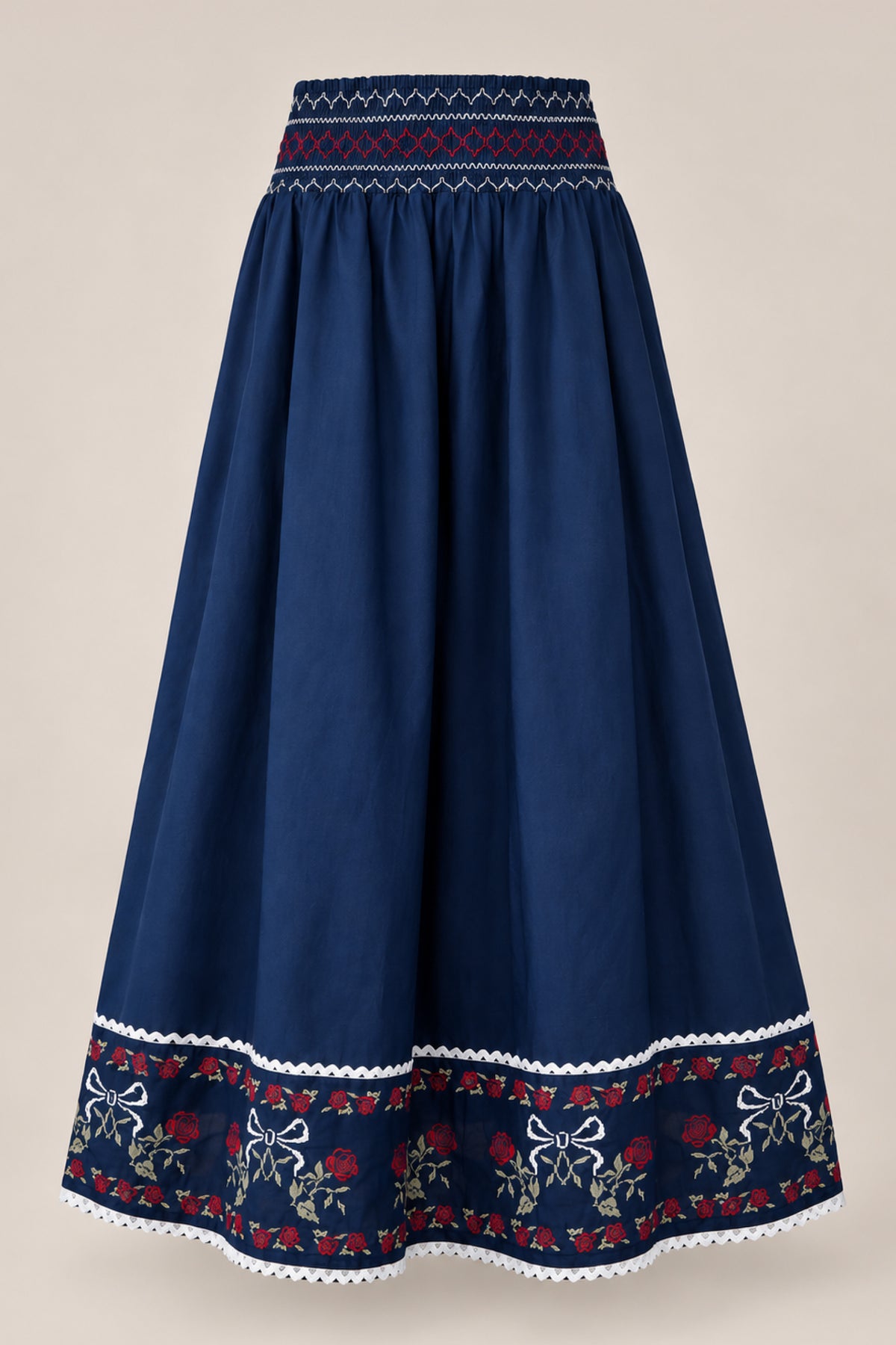 The Bluebell Skirt in Sapphire Rosette Embroidery by Ivy City Co is a long, navy blue A-line cotton skirt featuring a patterned waistband, intricate floral embroidery, and charming white bows near the hem. Delicate white lace trim adorns the bottom edge, with the skirt displayed on a plain background.