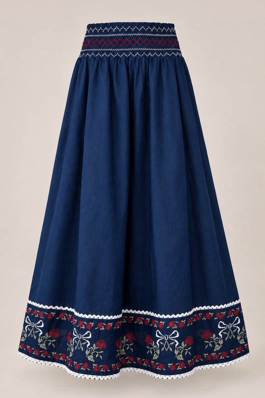 The Bluebell Skirt in Sapphire Rosette Embroidery by Ivy City Co is a long, navy blue A-line cotton skirt featuring a patterned waistband, intricate floral embroidery, and charming white bows near the hem. Delicate white lace trim adorns the bottom edge, with the skirt displayed on a plain background.