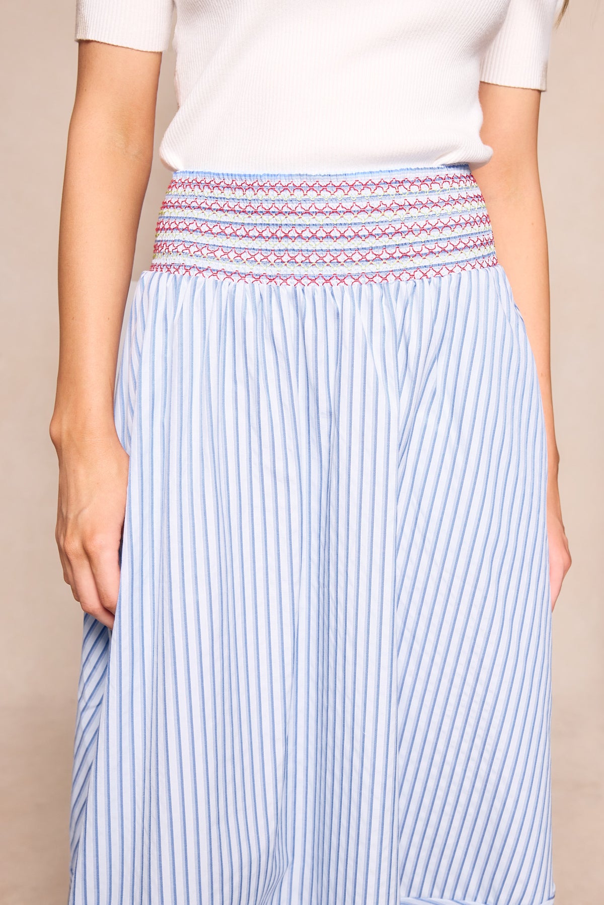 A person wears a white short-sleeve top and the Ivy City Co Bluebell Skirt in Blue & White Stripes, featuring a pink and blue embroidered waistband. Only the torso and upper legs are visible. This look is also maternity friendly.